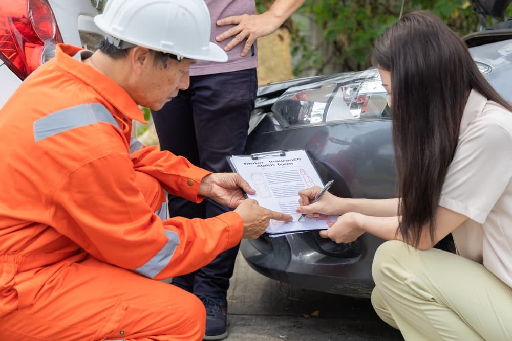 Inspector documenting details on clipboard near damaged car while drivers exchange insurance information.