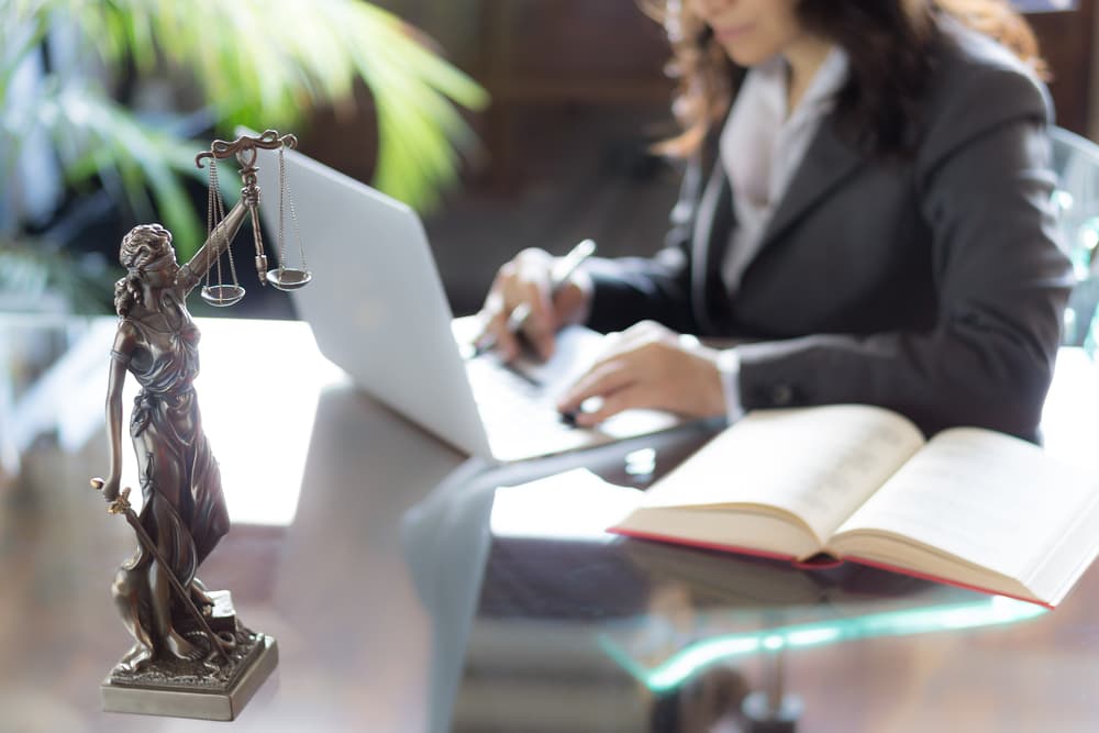 Statue of Justice with scales in lawyer's office, attorney working on laptop. Legal advice concept.