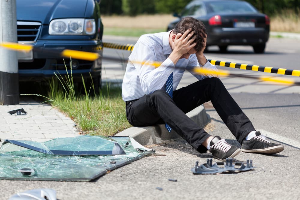 A distressed man sits by shattered glass and a car after an accident.